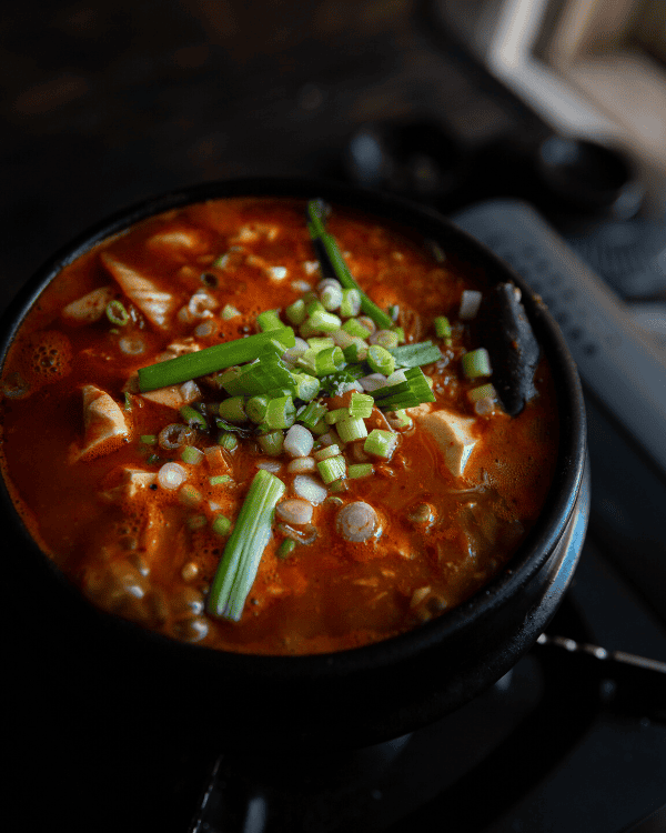 A bowl of spicy Korean tofu stew (soondubu jjigae) filled with soft tofu, green onions, and vegetables, all in a rich red broth. The dish is served in a traditional black stone pot.