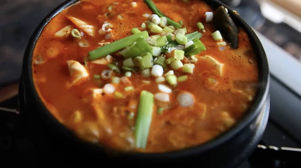 A steaming bowl of spicy Korean soondooboo chigae, garnished with chopped green onions and vibrant kimchi, served in a traditional black stone pot.