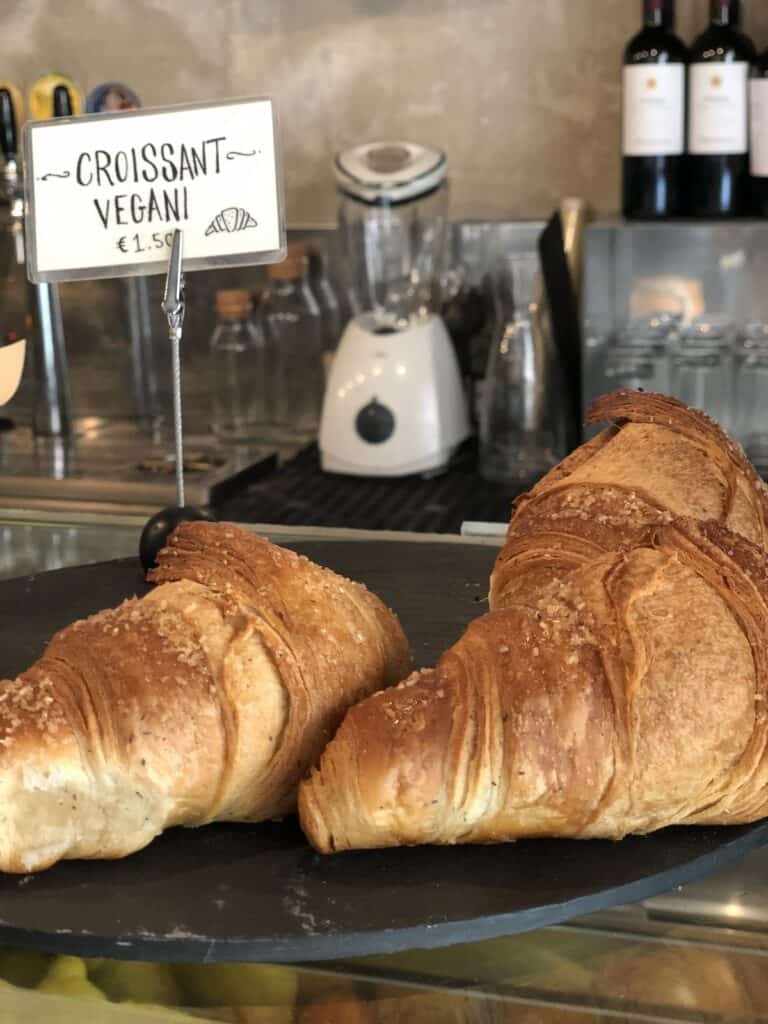 Traveling as a Vegan in Italy: An Easy & Delicious Guide. 11 Two large vegan croissants displayed on a round black tray, labeled CROISSANT VEGANI with a price of €1.50. A blender and wine bottles are visible in the background, evoking a charming Italian café ambiance.