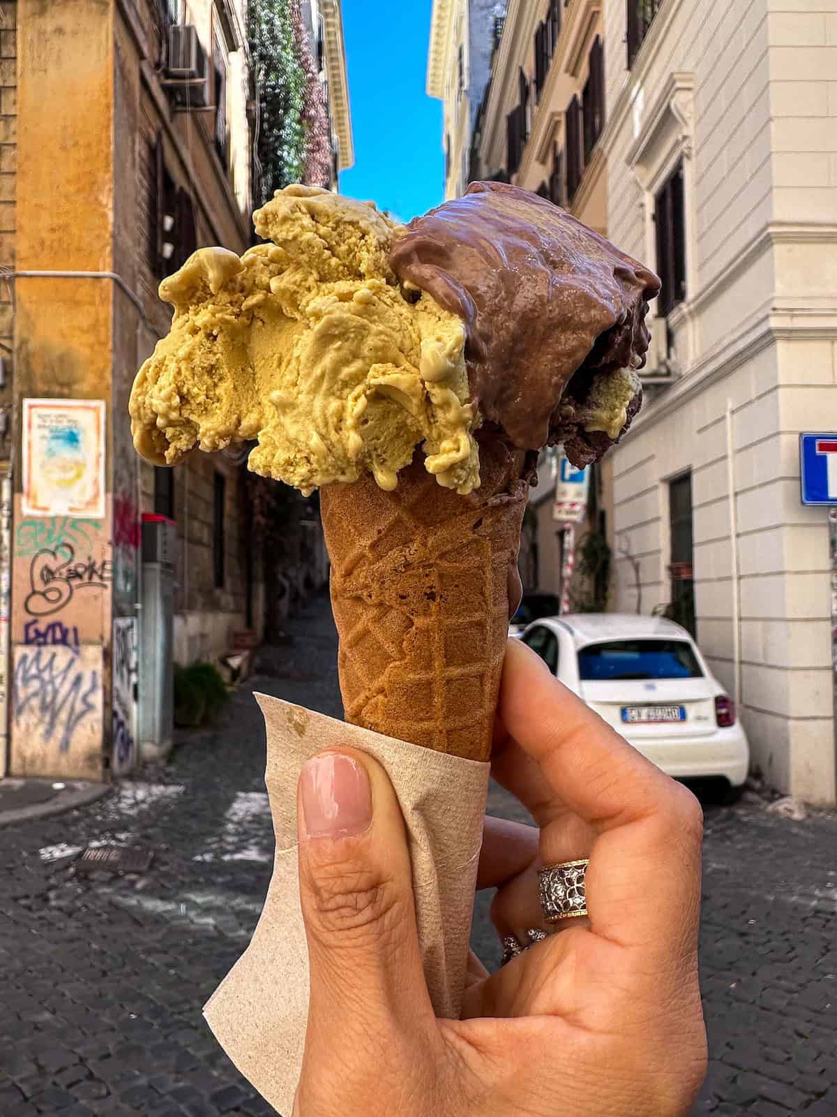 Traveling as a Vegan in Italy: An Easy & Delicious Guide. 13 A person holds a waffle cone with two scoops of gelato—pistachio and chocolate—against a narrow, quaint Roman street in Italy, with cobblestones, parked cars, and buildings adorned with street art and ivy-covered walls.