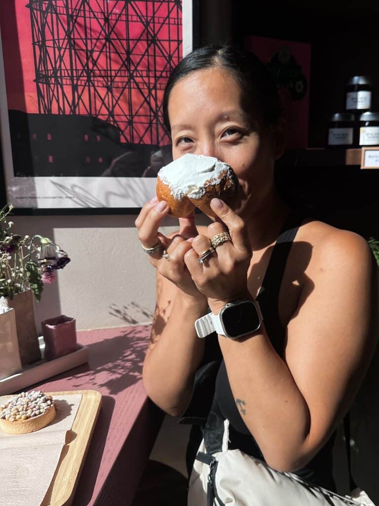 Traveling as a Vegan in Italy: An Easy & Delicious Guide. 12 Joanne holding a frosted vegan pastry near her face, smiling. She is in a room with an Italian flair, featuring a pink table, a poster, and sunlight streaming through the window. Another sprinkle-adorned pastry rests nearby on the table.