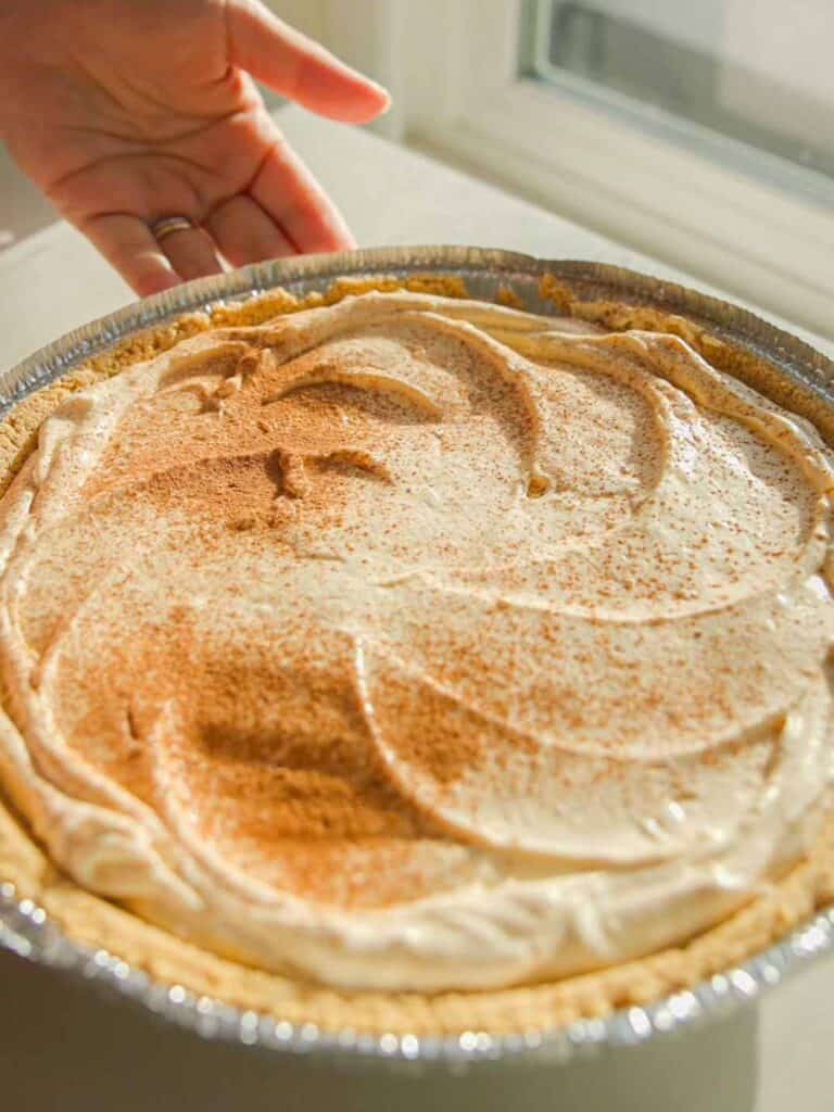 6-Ingredient BEST No-Bake Pumpkin Cheesecake (Vegan). 11 A hand holding a pie with a golden crust and creamy filling sprinkled with cinnamon, presented in a foil pie tin, set against a softly lit window background.