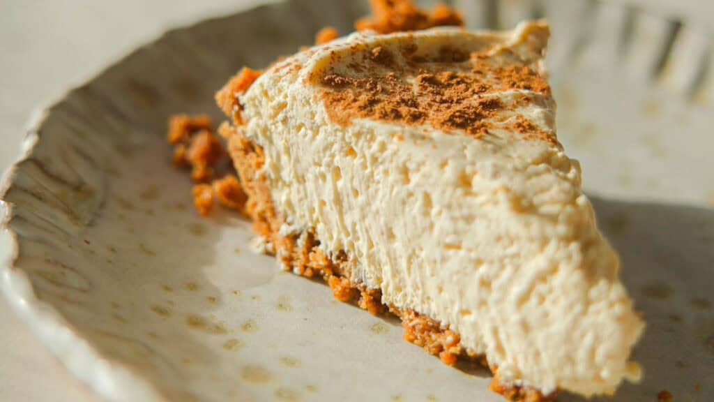 6-Ingredient BEST No-Bake Pumpkin Cheesecake (Vegan). 1 A slice of creamy no-bake pumpkin cheesecake with a graham cracker crust is placed on a decorative ceramic plate. The cheesecake is topped with a dusting of cinnamon, and the image is warmly lit to highlight its texture.