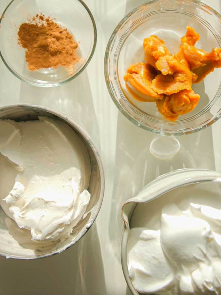 6-Ingredient BEST No-Bake Pumpkin Cheesecake (Vegan). 3 Four bowls on a light table containing the ingredients for this vegan no-bake pumpkin cheesecake: one with cinnamon powder, another with pumpkin puree, a third with whipped cream, and the last with cream cheese. The sunlight casts soft shadows over the ingredients.