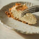 30+ AMAZING VEGAN Holiday Recipes Everyone's gonna Love. 32 A slice of creamy pumpkin cheesecake with a crumbly graham cracker crust rests on a textured plate. A fork is cutting into the cake, casting a soft shadow across the plate. The scene is warmly lit, highlighting the cake's smooth texture.