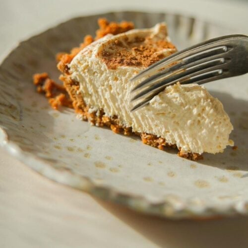 6-Ingredient BEST No-Bake Pumpkin Cheesecake (Vegan). 17 A slice of creamy pumpkin cheesecake with a crumbly graham cracker crust rests on a textured plate. A fork is cutting into the cake, casting a soft shadow across the plate. The scene is warmly lit, highlighting the cake's smooth texture.