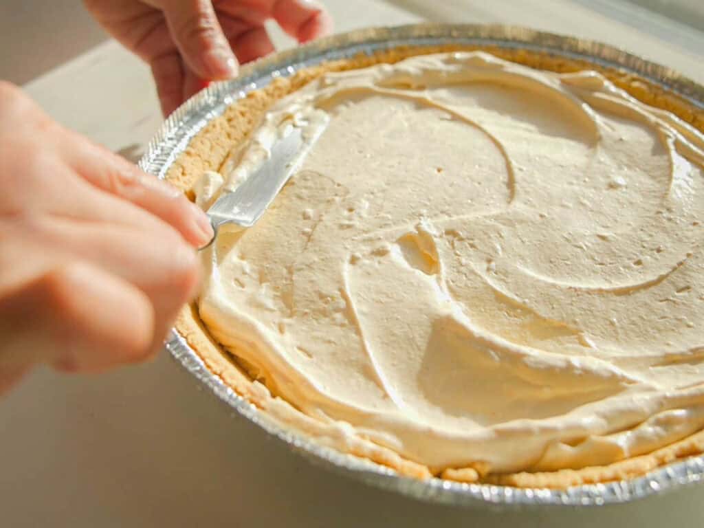 6-Ingredient BEST No-Bake Pumpkin Cheesecake (Vegan). 10 A person spreads creamy filling over a pie crust in a foil dish with a knife. The pie is on a wooden surface, and the sunlight casts a warm glow over the scene.