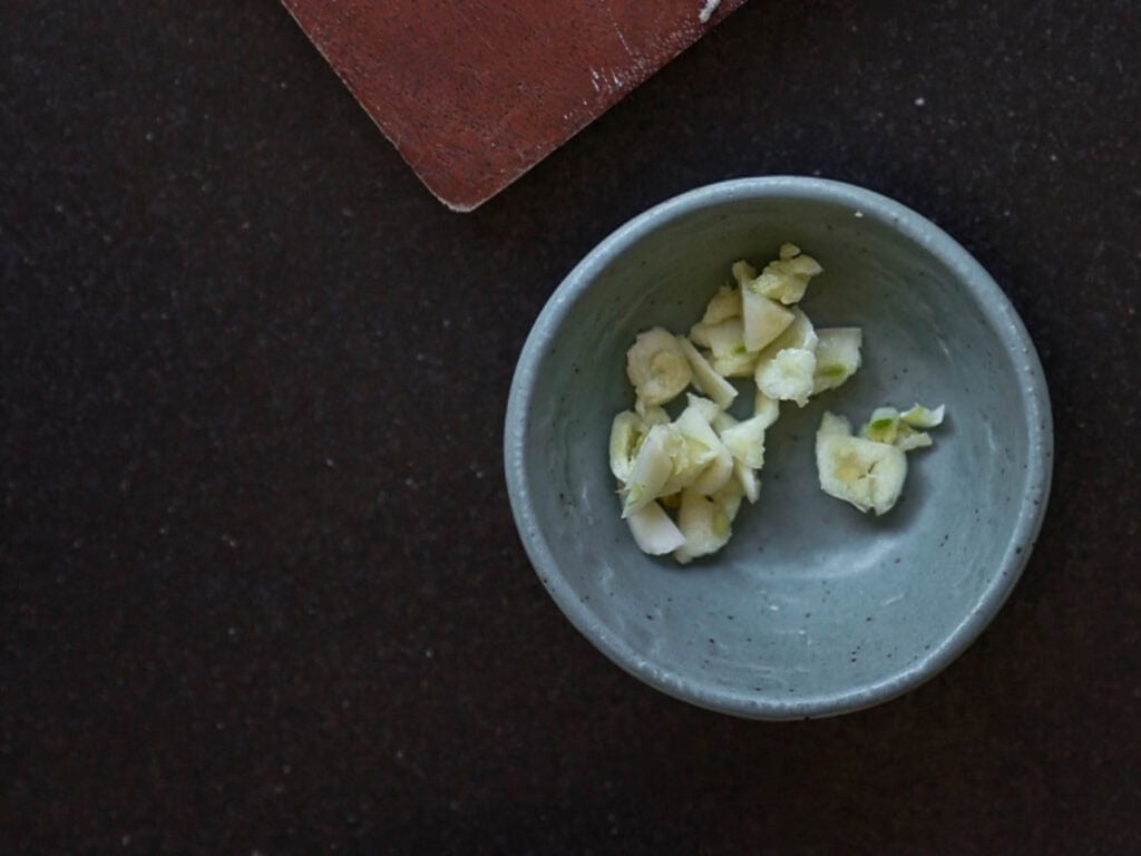 5-Ingredient Easy Chickpea Pasta Recipe (Pasta e Ceci). 5 A small green bowl filled with chopped garlic pieces is placed on a dark countertop. A wooden board is partially visible in the top left corner.