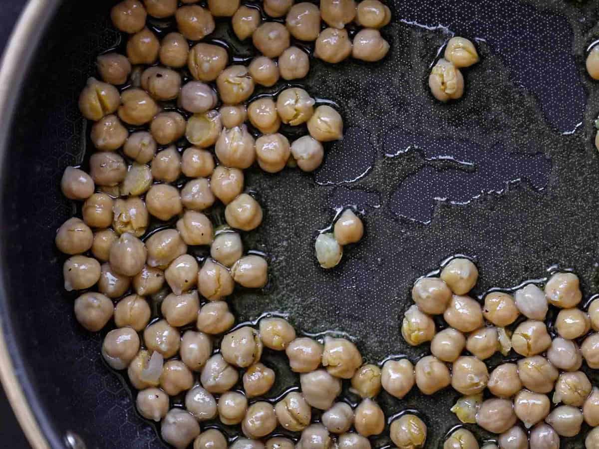 5-Ingredient Easy Chickpea Pasta Recipe (Pasta e Ceci). 7 Chickpeas being sautéed in a black frying pan with a small amount of oil. The chickpeas are spread out, and the pans surface is visible.