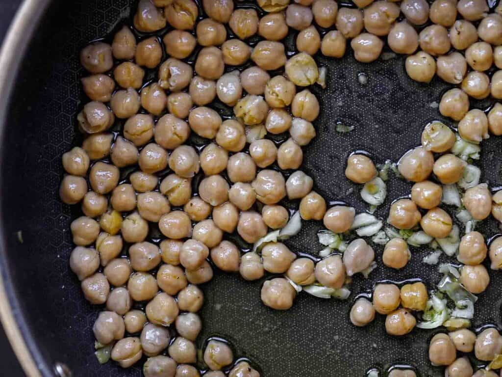 5-Ingredient Easy Chickpea Pasta Recipe (Pasta e Ceci). 4 Chickpeas and chopped garlic cooking in a black pan with oil, a close-up view showing the chickpeas evenly distributed and glistening in the light.