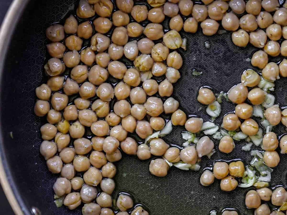 5-Ingredient Easy Chickpea Pasta Recipe (Pasta e Ceci). 8 Chickpeas and chopped garlic cooking in a black pan with oil, a close-up view showing the chickpeas evenly distributed and glistening in the light.