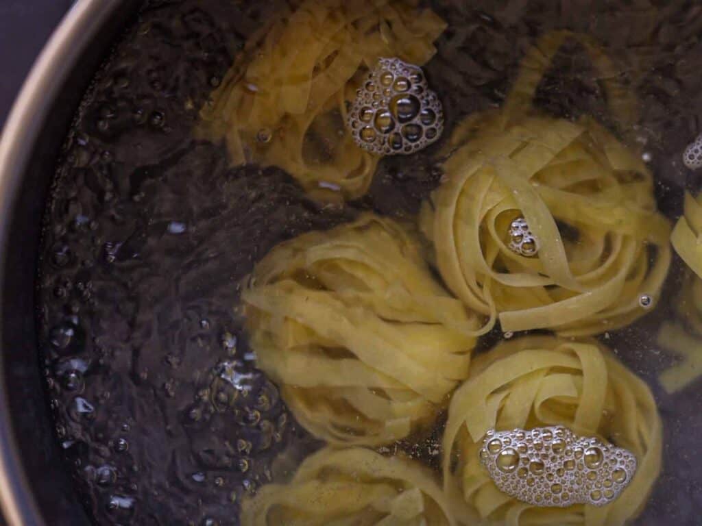 5-Ingredient Easy Chickpea Pasta Recipe (Pasta e Ceci). 3 Tagliatelle pasta nests boiling in water, with bubbles forming around them in a pot.