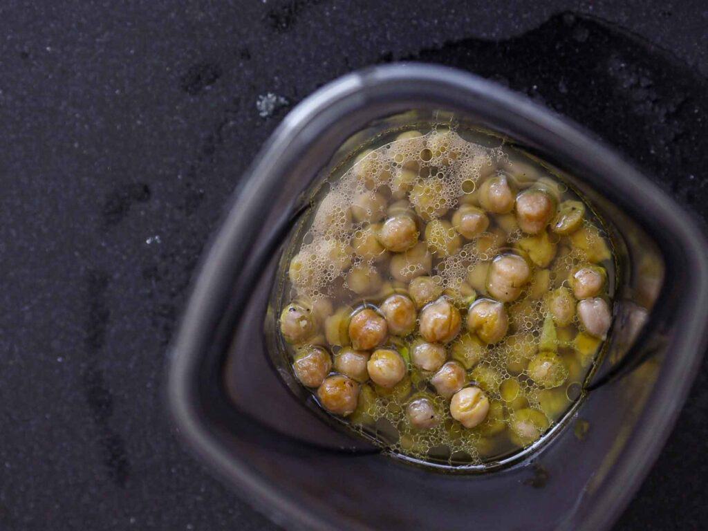 5-Ingredient Easy Chickpea Pasta Recipe (Pasta e Ceci). 11 A top view of a blender container filled with soaked chickpeas and their cooking liquid. The chickpeas are partially visible through the transparent sides, with bubbles forming on the surface of the liquid. The container is on a dark countertop.