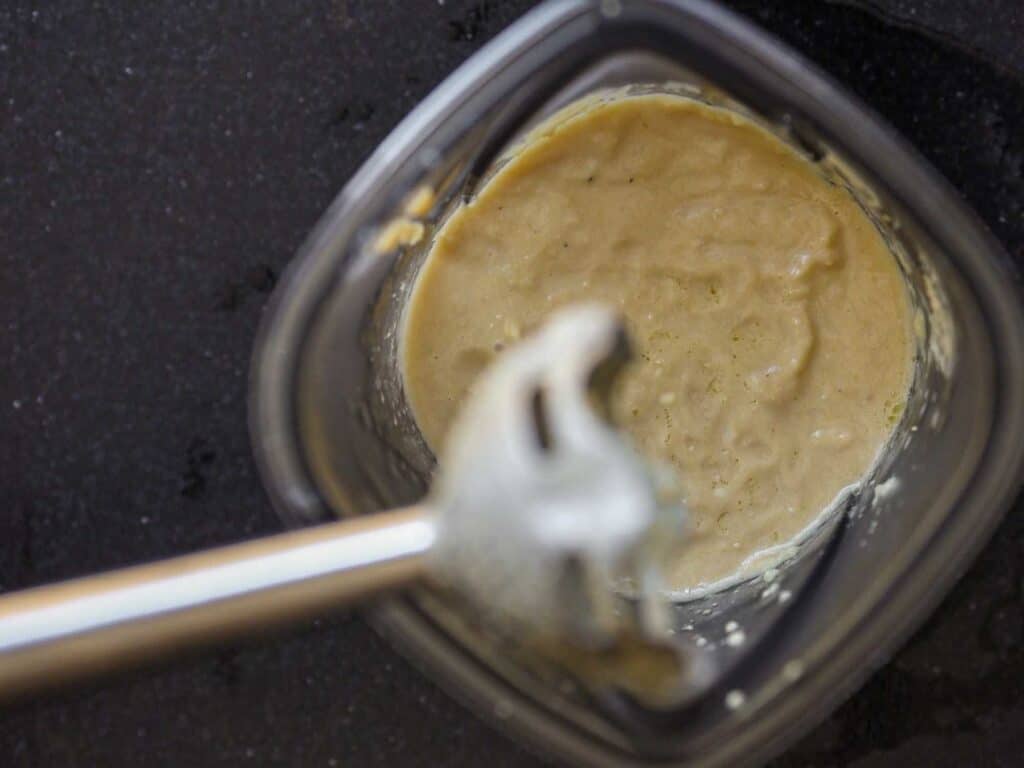 5-Ingredient Easy Chickpea Pasta Recipe (Pasta e Ceci). 12 Close-up of a blending container with a creamy light brown mixture inside. An immersion blender, partially submerged, is positioned in the center. The container rests on a dark countertop.