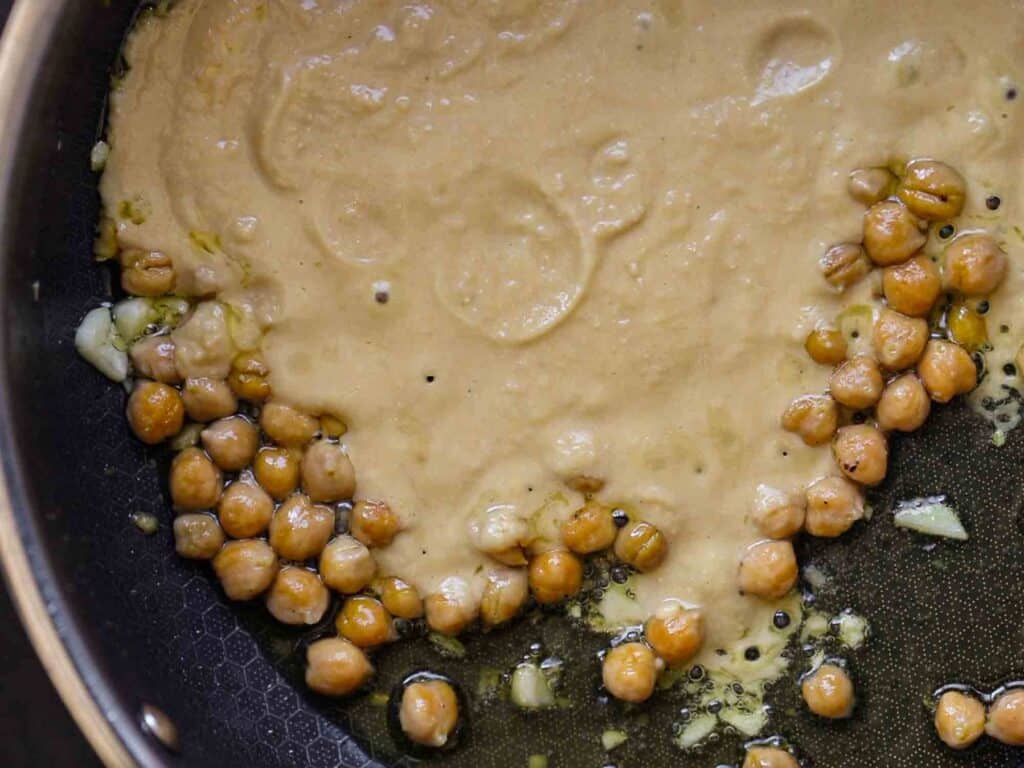 5-Ingredient Easy Chickpea Pasta Recipe (Pasta e Ceci). 14 Chickpeas and a creamy sauce cooking in a black skillet with bits of minced garlic. The chickpeas are partially covered by the sauce, which is light brown and smooth in texture.