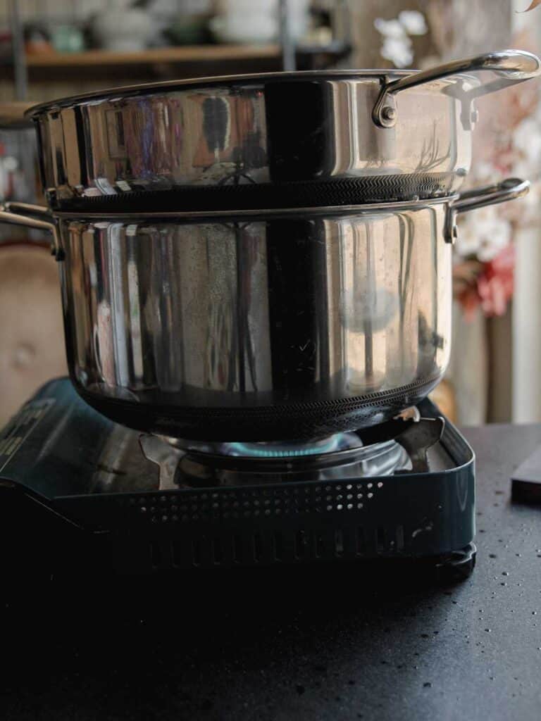 5-Ingredient Easy Chickpea Pasta Recipe (Pasta e Ceci). 9 Two stacked stainless steel pots are placed on a portable gas burner. The burner flame is visible, and the background shows blurred kitchen items on shelves.
