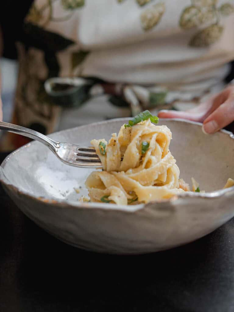5-Ingredient Easy Chickpea Pasta Recipe (Pasta e Ceci). 2 A person twirls fettuccine pasta with a fork in a textured ceramic bowl. The pasta is garnished with fresh green herbs and grated cheese. The background shows the persons torso wearing an apron.