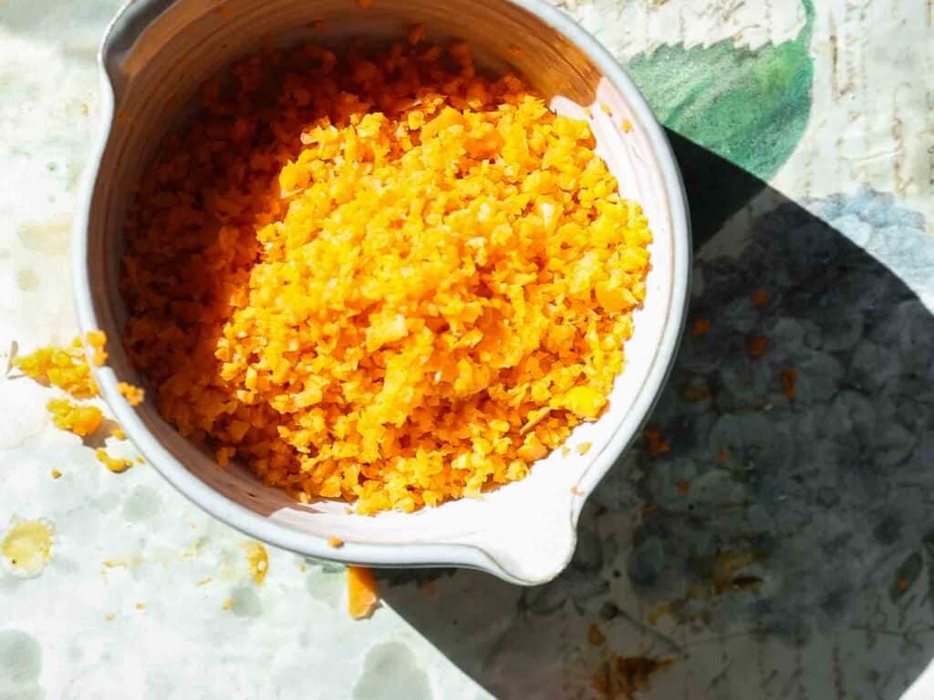 The BEST Vegan Carrot Cake Recipe - Not Too Sweet! 3 Close-up of a bowl filled with finely grated carrot, placed on a patterned surface. Sunlight creates shadows, highlighting the vibrant color and texture of the zest.