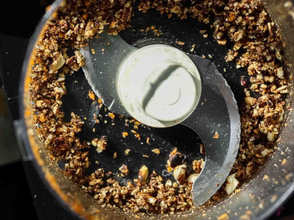 The BEST Vegan Carrot Cake Recipe - Not Too Sweet! 11 Close-up of a food processor with chopped nuts and ingredients around the central blade, showing a mix of brown and beige colors and a textured appearance.