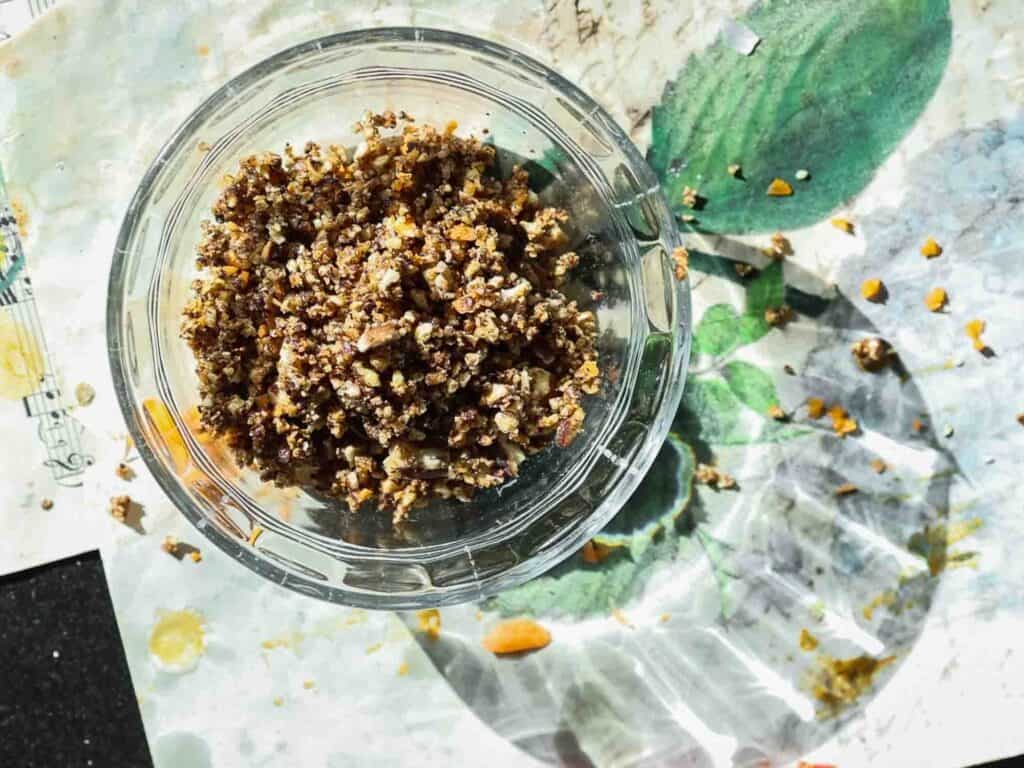 The BEST Vegan Carrot Cake Recipe - Not Too Sweet! 8 A clear glass bowl filled with a mixture of chopped nuts on a patterned surface. Some crumbs are scattered around the bowl, catching the sunlight.