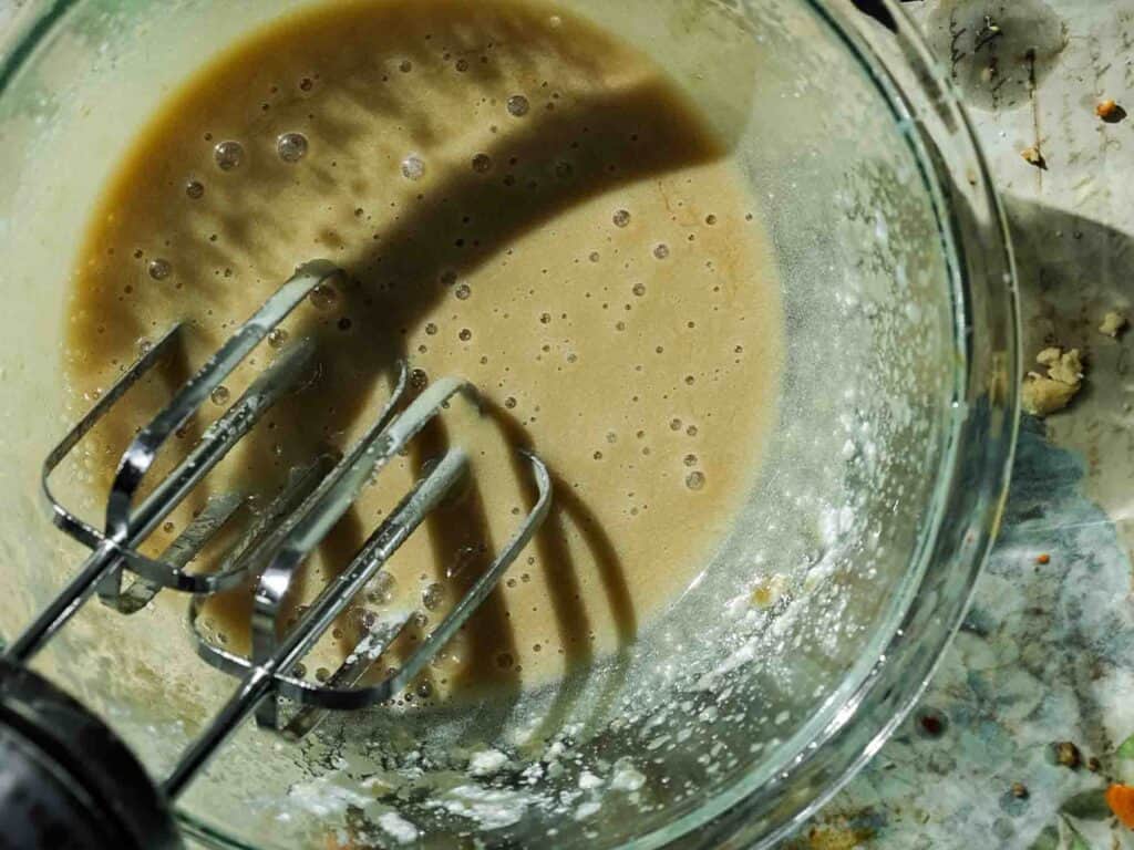The BEST Vegan Carrot Cake Recipe - Not Too Sweet! 16 A glass bowl containing tan batter is shown from above, partially mixed with an electric mixer with two beaters. The bowl rests on a speckled surface, and light casts a shadow of the beaters onto the batter.