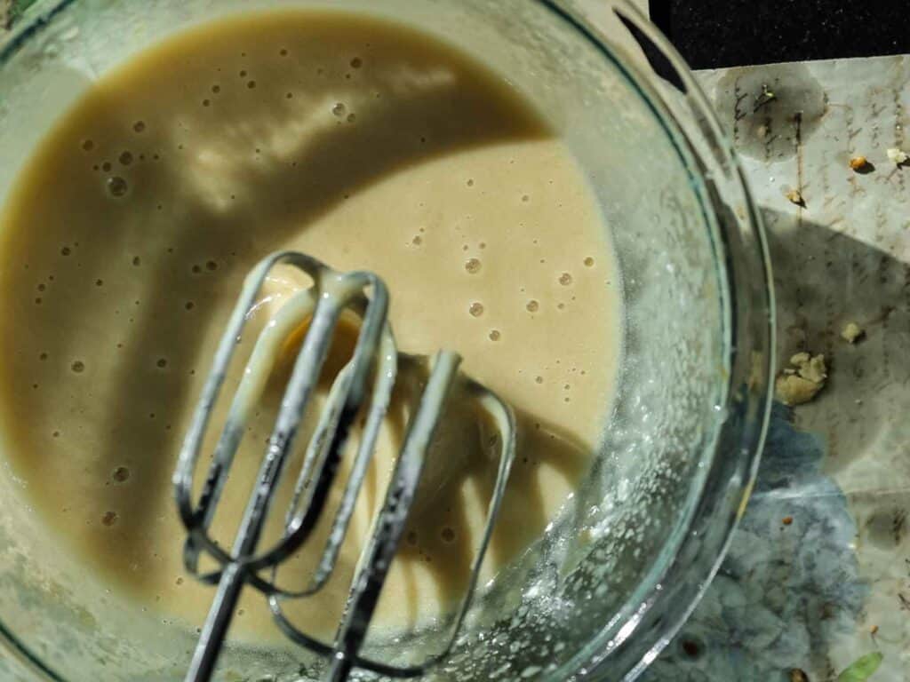 The BEST Vegan Carrot Cake Recipe - Not Too Sweet! 17 A glass bowl containing a creamy batter with air bubbles is shown. Two metal beaters are partially submerged in the mixture, resting against the side of the bowl. A textured, slightly messy surface is visible beside the bowl.