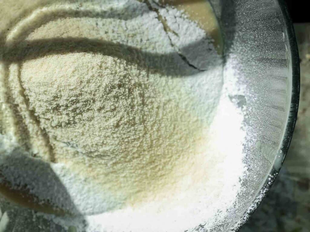 The BEST Vegan Carrot Cake Recipe - Not Too Sweet! 14 A close-up view of white powdery flour in a clear glass mixing bowl. Natural light casts soft shadows across the textured surface, highlighting its smooth, granular appearance.