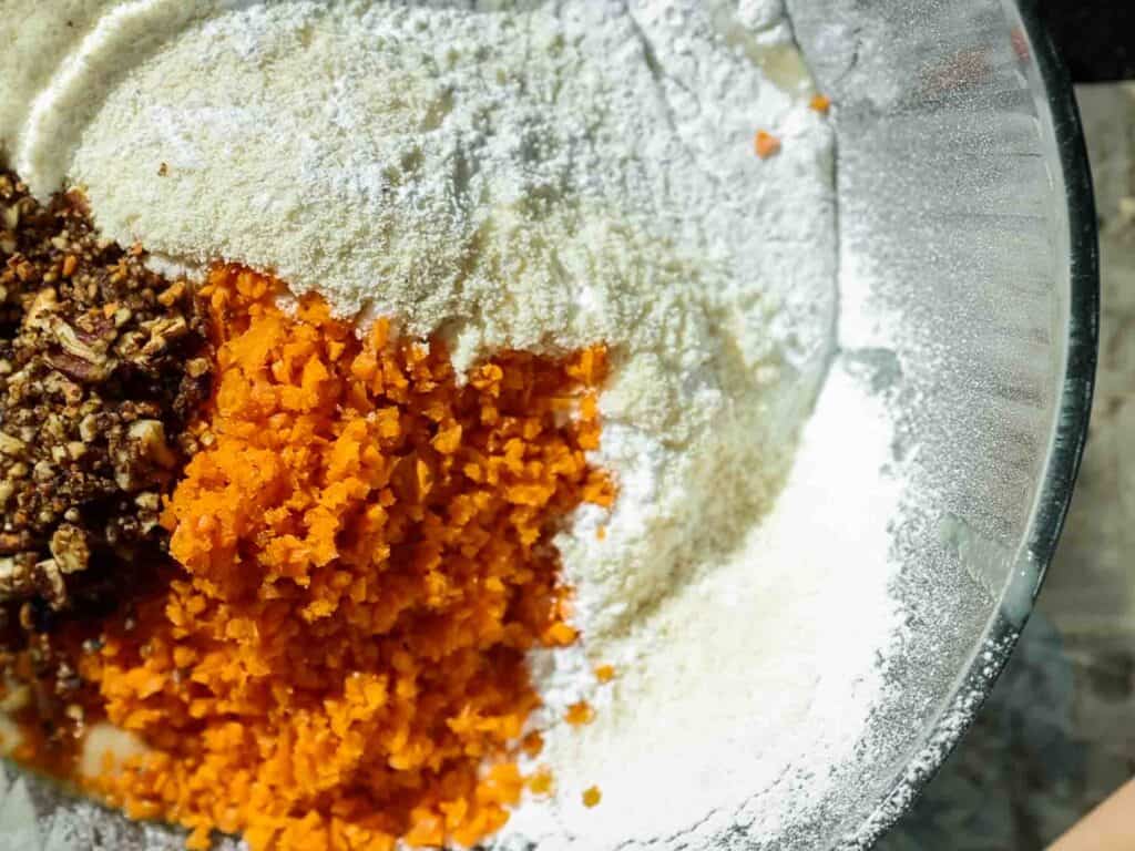 The BEST Vegan Carrot Cake Recipe - Not Too Sweet! 18 A clear glass bowl containing flour, grated carrots, and chopped nuts, ready for mixing. The ingredients are positioned on a textured surface, capturing the shadows and highlights.