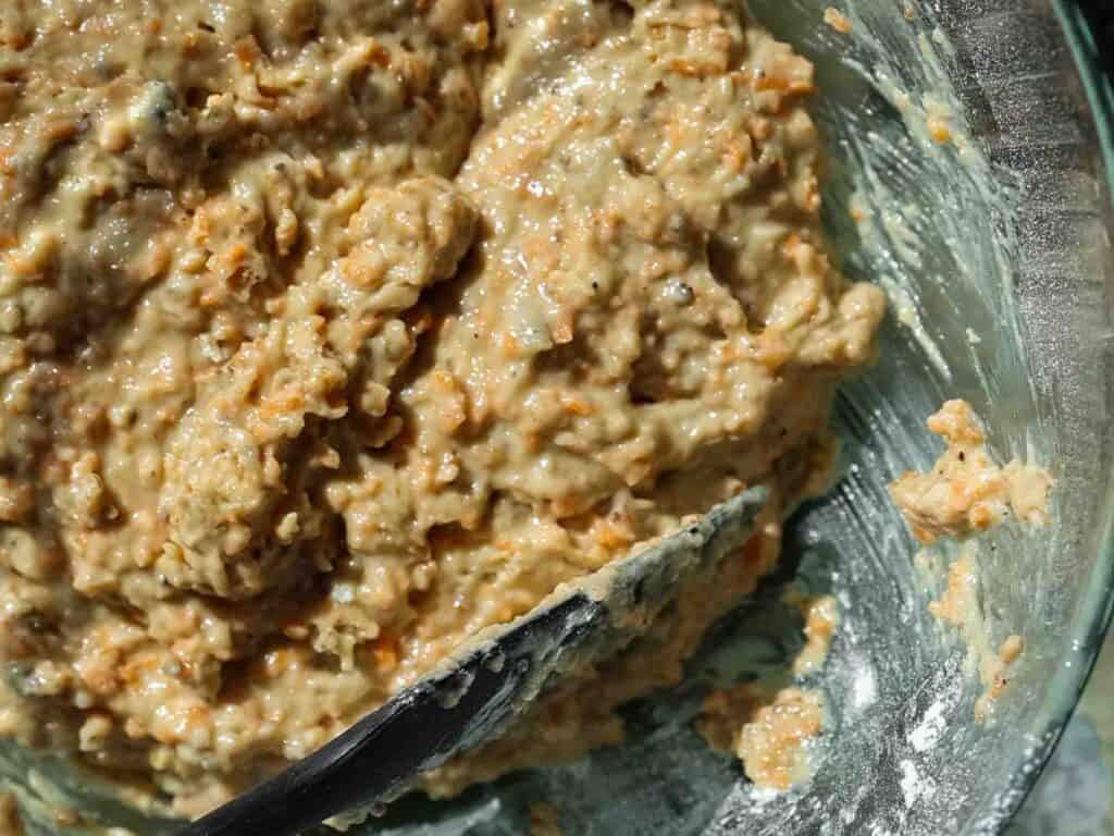 The BEST Vegan Carrot Cake Recipe - Not Too Sweet! 19 Close-up of a bowl containing thick, light brown vegan carrot cake batter with small visible bits of grated carrot. A mixing spoon is partially visible, coated with the batter. The glass bowl shows some flour residue on its side.