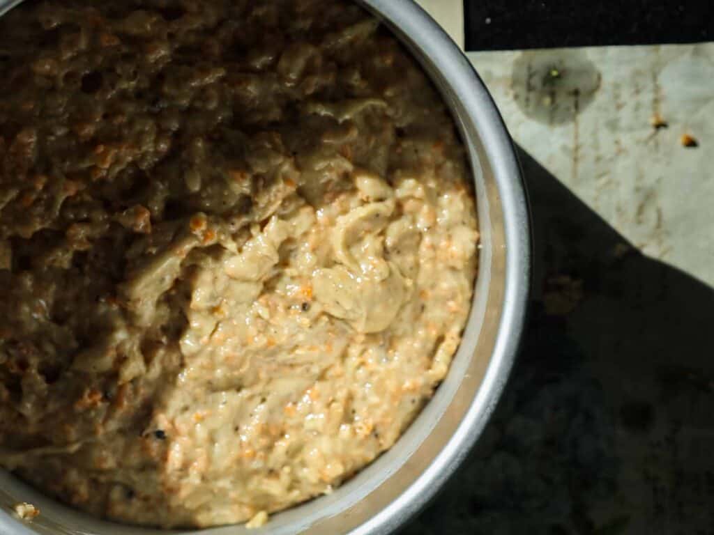 The BEST Vegan Carrot Cake Recipe - Not Too Sweet! 20 A close-up of a vegan carrot cake batter in round cake pan. The mixture appears to have a light brown color with small dark flecks throughout and is placed on a surface with shadows visible in the background.