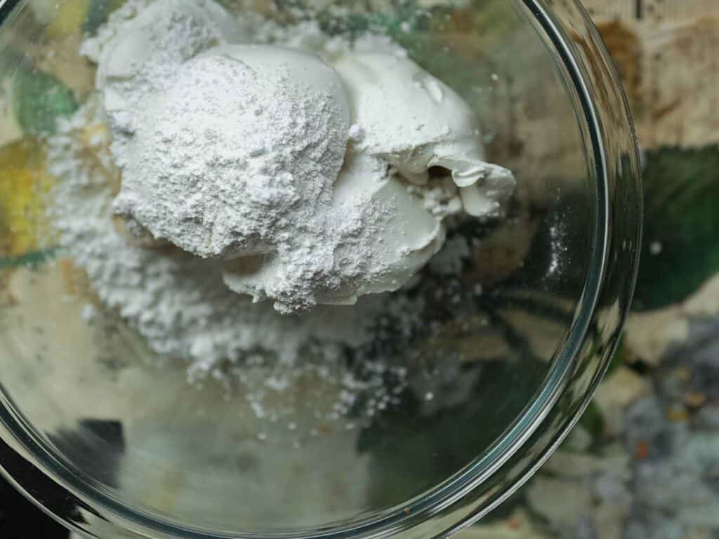 The BEST Vegan Carrot Cake Recipe - Not Too Sweet! 9 A clear glass bowl containing heaped tablespoons of white powdery substance and creamy mixture, set on a blurred background with hints of green and yellow.