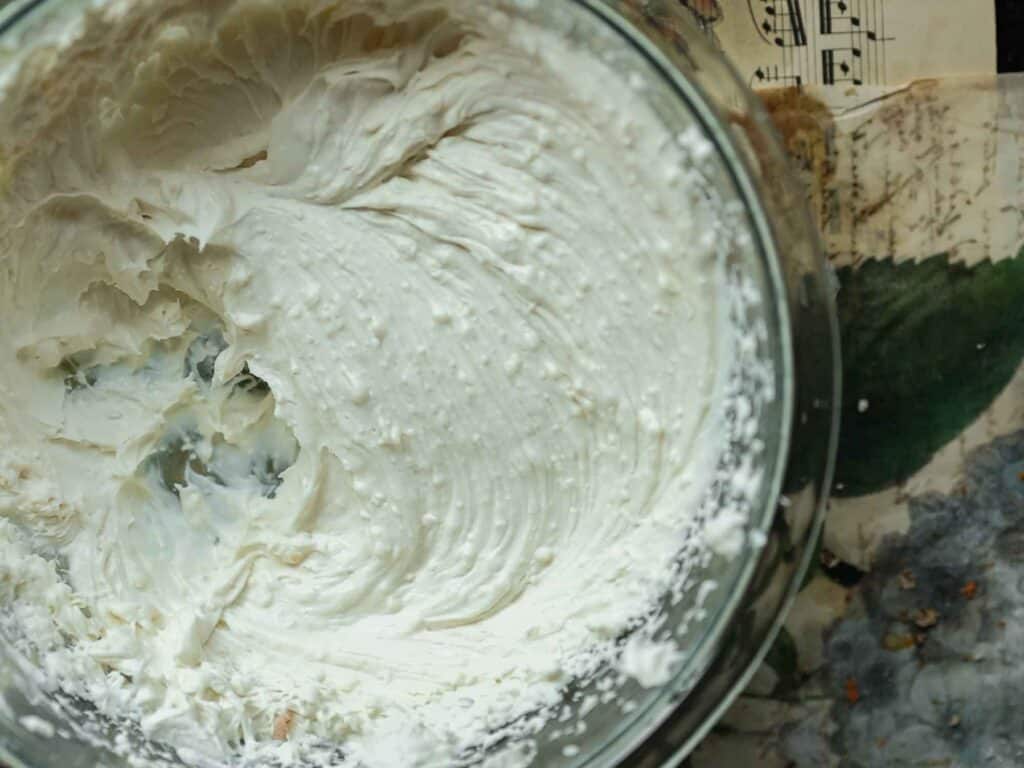 The BEST Vegan Carrot Cake Recipe - Not Too Sweet! 23 A glass bowl filled with whipped vegan cream sits on a floral and music sheet background. The cream is smooth, with some uneven textures and swirls, showing signs of being recently mixed or whipped.