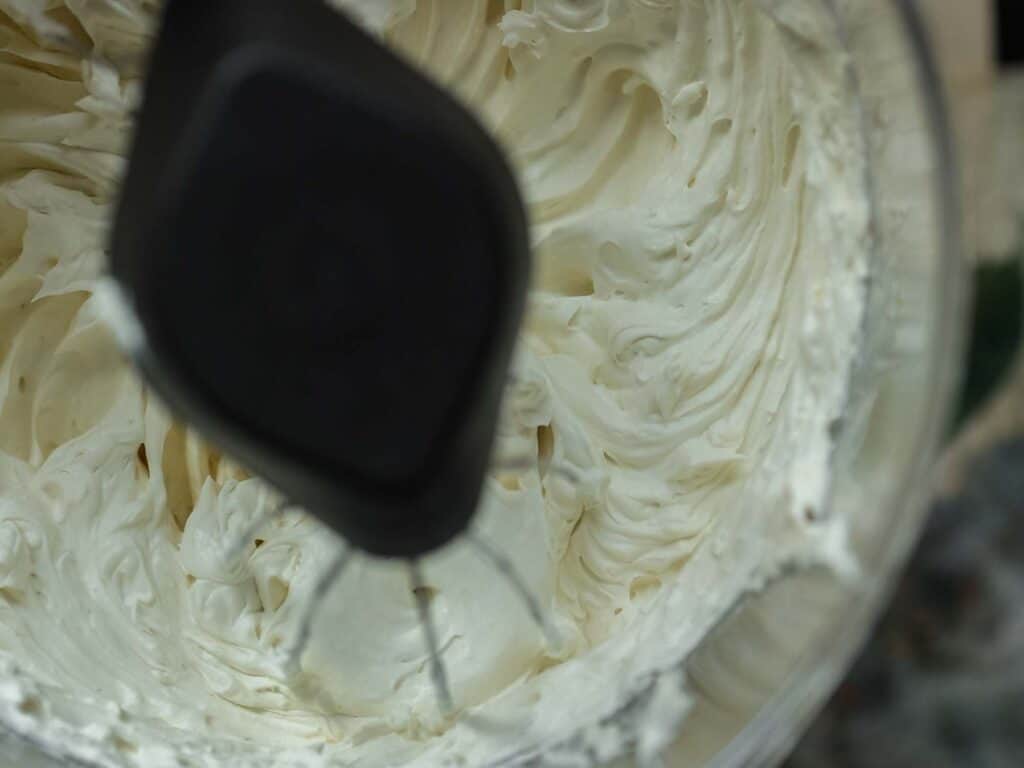 The BEST Vegan Carrot Cake Recipe - Not Too Sweet! 10 Close-up of a mixer blending smooth, creamy vegan frosting in a bowl. The spiraled texture of the frosting is visible, with a beater attachment partially inserted. The creamy color contrasts with the dark handle of the mixer.