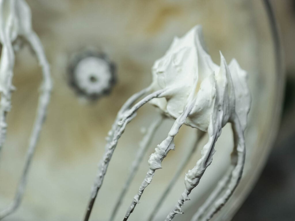 The BEST Vegan Carrot Cake Recipe - Not Too Sweet! 25 Close-up of a mixer with two beaters covered in thick, whipped cream. The cream is smooth and fluffy, highlighting the mixer blades in a kitchen setting.
