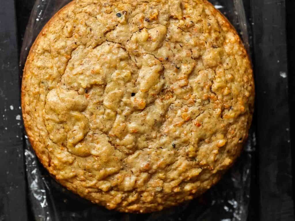 The BEST Vegan Carrot Cake Recipe - Not Too Sweet! 21 A close-up of a round, golden-brown vegan carrot cake with a textured surface, resting on a dark background.