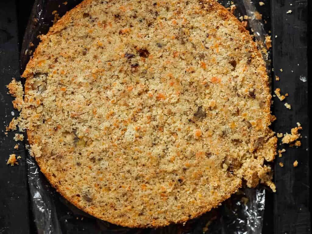 The BEST Vegan Carrot Cake Recipe - Not Too Sweet! 26 A close-up of a single-layered carrot cake on a dark surface. The cakes crumbly texture and visible bits of carrot and nuts are apparent. Some scattered crumbs surround the cake.