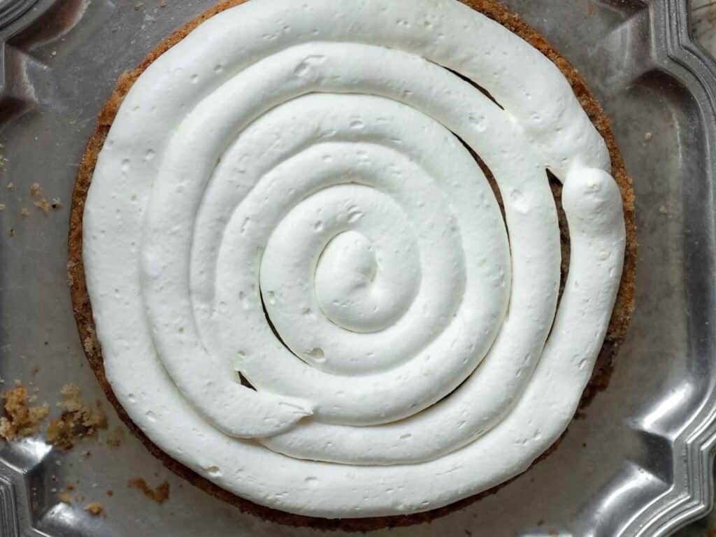 The BEST Vegan Carrot Cake Recipe - Not Too Sweet! 27 A round pastry with a swirl of white whipped cream on top, placed on a metal tray. Crumbs are visible around the edges.