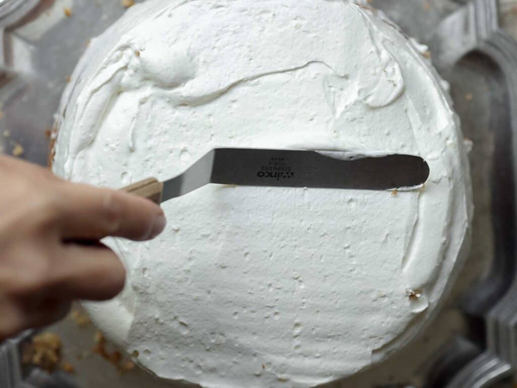 The BEST Vegan Carrot Cake Recipe - Not Too Sweet! 28 A hand is using a spatula to spread white icing over a round cake. The cake is on a clear surface, and crumbs are visible on the edges of the icing.