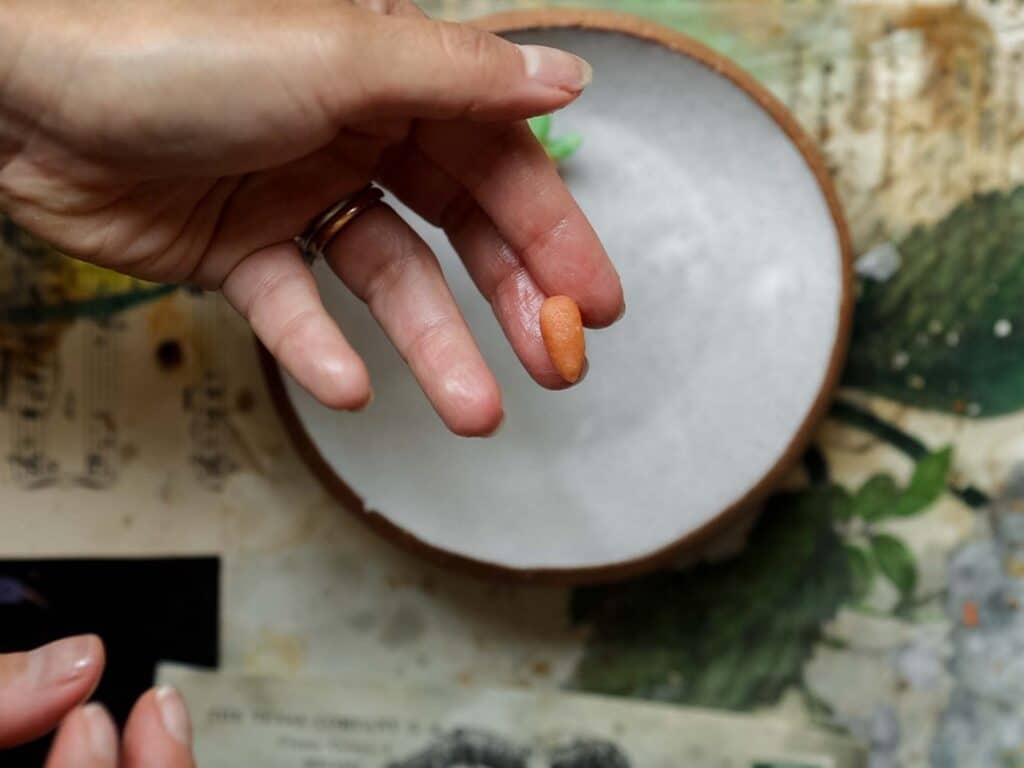 The BEST Vegan Carrot Cake Recipe - Not Too Sweet! 31 A hand is holding a small, oval-shaped bit or orange marzipan, between the thumb and index finger. In the background, there is a white bowl on a table with floral patterns and sheet music.