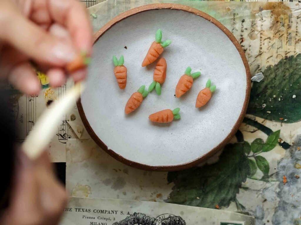 The BEST Vegan Carrot Cake Recipe - Not Too Sweet! 32 A person shaping small sculptures of carrots from marzipan or a similar material. Several finished carrot sculptures are placed on a round plate. The scene is set on a table with scattered papers and artistic supplies.