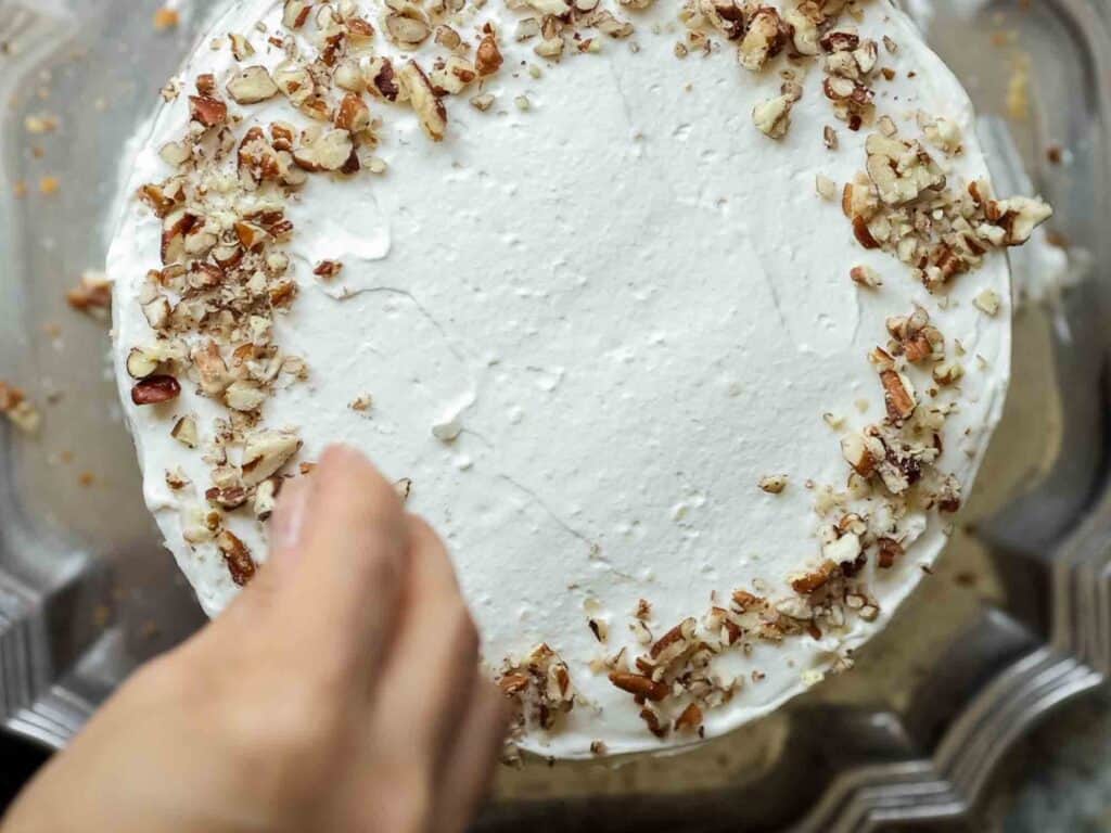 The BEST Vegan Carrot Cake Recipe - Not Too Sweet! 30 A hand sprinkles chopped nuts on top of a cake with smooth white frosting. The cakes surface is decorated with a ring of crushed nuts around the edges.