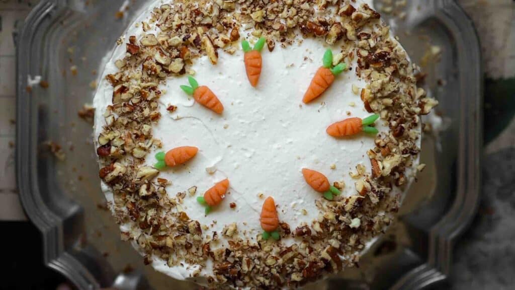 The BEST Vegan Carrot Cake Recipe - Not Too Sweet! 1 A round vegan carrot cake topped with white frosting, adorned with small fondant carrots in a circle on top. Chopped nuts line the edges, adding texture and contrast to the smooth icing.