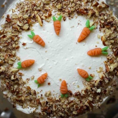 The BEST Vegan Carrot Cake Recipe - Not Too Sweet! 38 A round vegan carrot cake topped with white frosting, adorned with small fondant carrots in a circle on top. Chopped nuts line the edges, adding texture and contrast to the smooth icing.