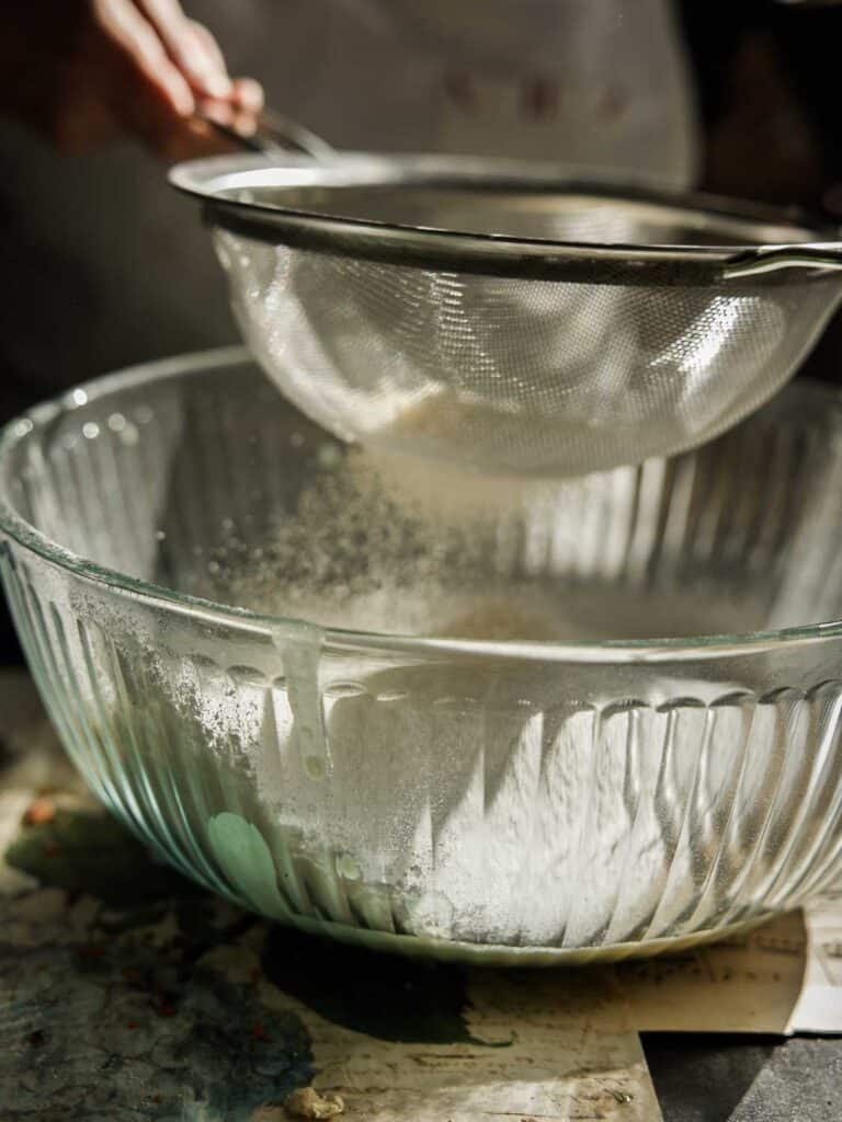 The BEST Vegan Carrot Cake Recipe - Not Too Sweet! 13 A person is sifting flour into a large glass bowl using a metal sieve. The flour is falling through the sieve, creating a light dust cloud in the bowl. The scene is warmly lit, highlighting the baking process.