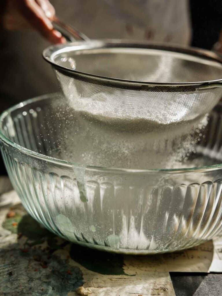 The BEST Vegan Carrot Cake Recipe - Not Too Sweet! 5 A person sifting flour through a metal sieve into a clear glass bowl. The scene is warmly lit, highlighting the falling flour and its particles. The countertop beneath appears rustic with a slight artistic touch.