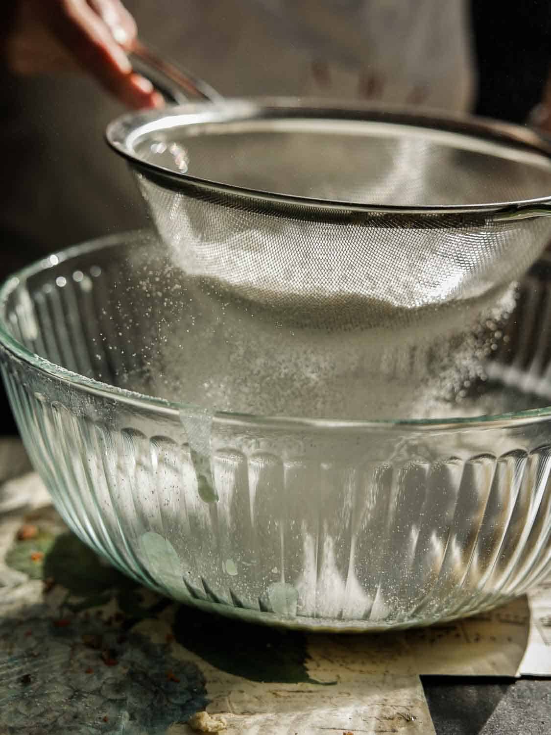The BEST Vegan Carrot Cake Recipe - Not Too Sweet! 12 A person sifting flour through a metal sieve into a clear glass bowl. The scene is warmly lit, highlighting the falling flour and its particles. The countertop beneath appears rustic with a slight artistic touch.