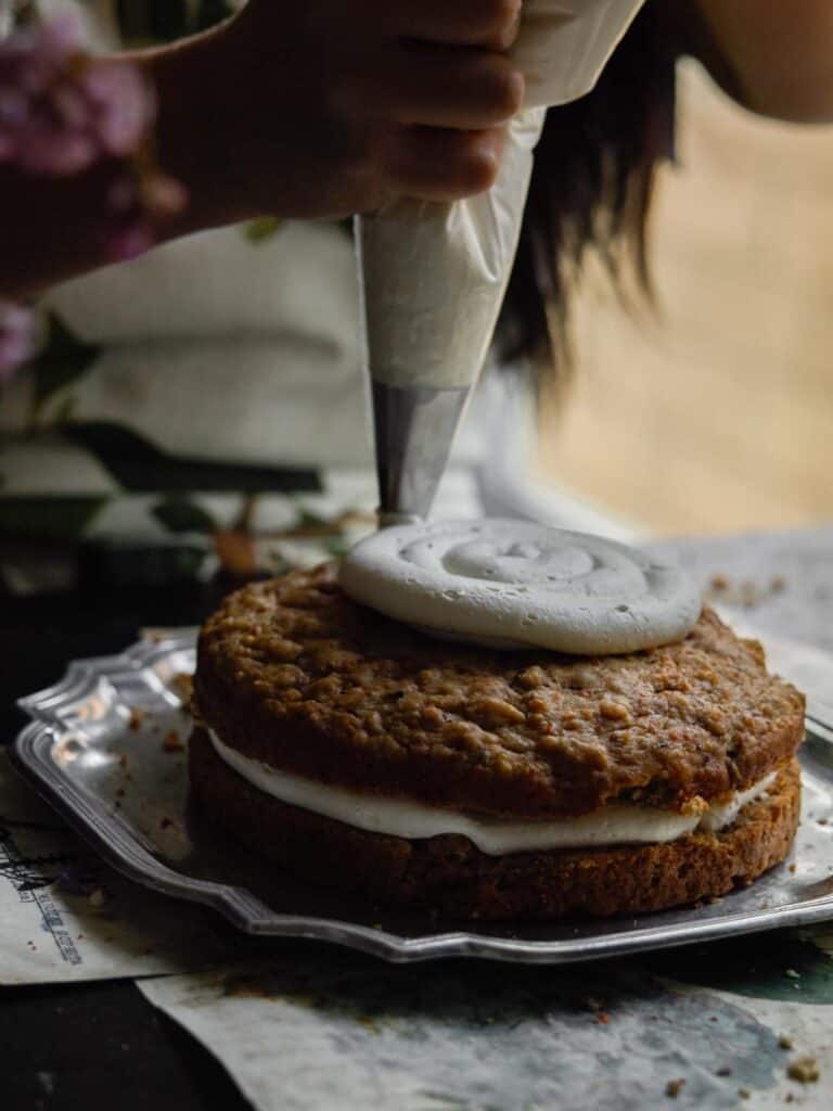 The BEST Vegan Carrot Cake Recipe - Not Too Sweet! 29 A person frosting a layer cake with white icing using a piping bag. The cake sits on a decorative silver tray, with an additional layer of icing between the two stacked cake layers. The scene is dimly lit, creating a cozy ambiance.