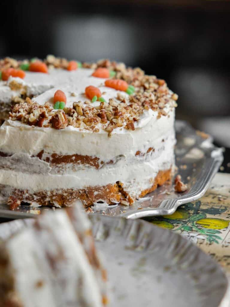 The BEST Vegan Carrot Cake Recipe - Not Too Sweet! 33 A layered vegan carrot cake topped with white frosting, chopped nuts, and small decorative fondant carrots on a silver platter. A slice is cut and placed on a nearby plate.