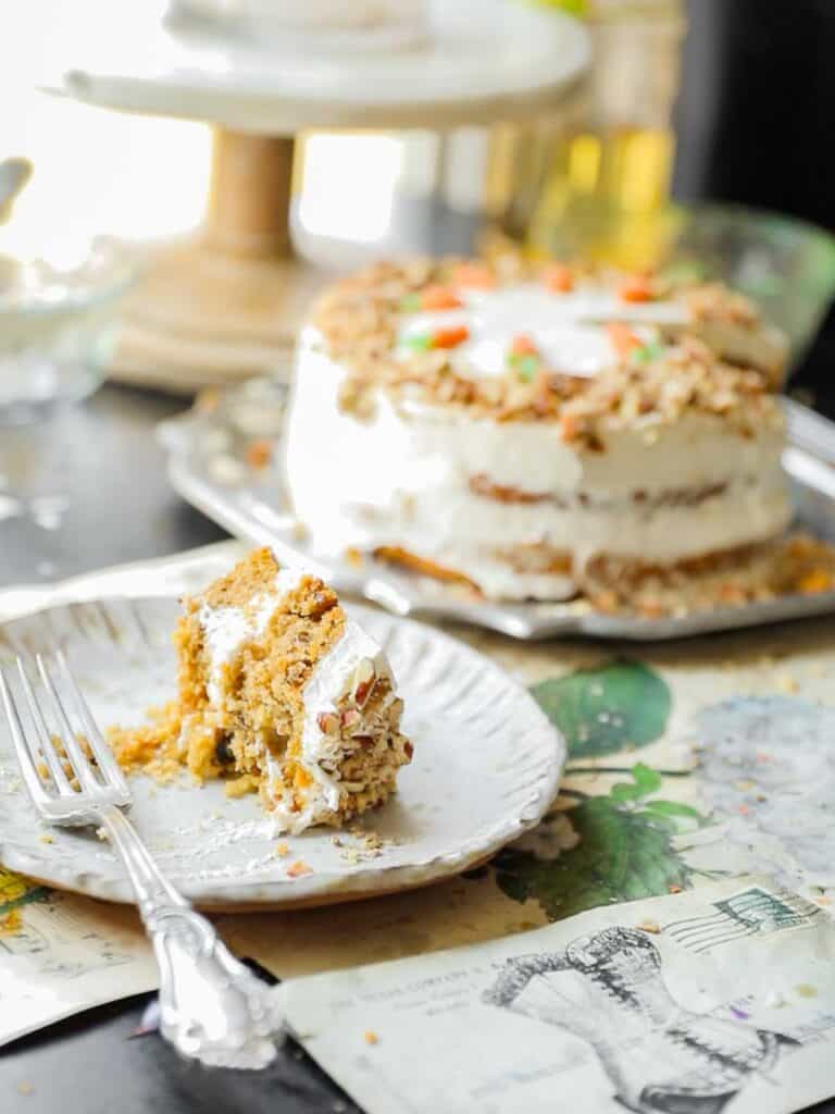 The BEST Vegan Carrot Cake Recipe - Not Too Sweet! 2 A slice of vegan carrot cake with cream cheese frosting rests on a white plate with a fork. The remaining cake is in the background, topped with frosting and walnuts. The table is decorated with illustrated papers.