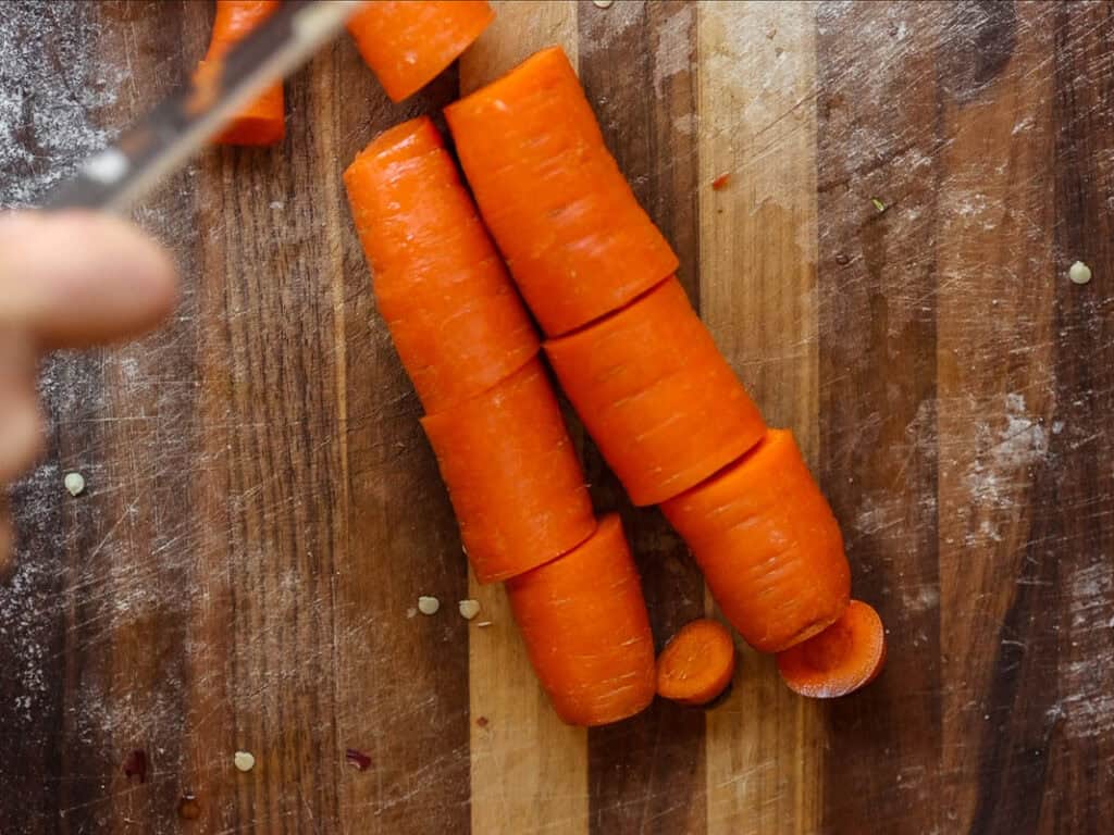 Gochujang and Roasted Carrot Soup - SPICY AND EASY. 3 Close-up of chopped orange carrots on a wooden cutting board. A hand is holding a knife in the top left corner, mid-action. Small carrot pieces are scattered around.